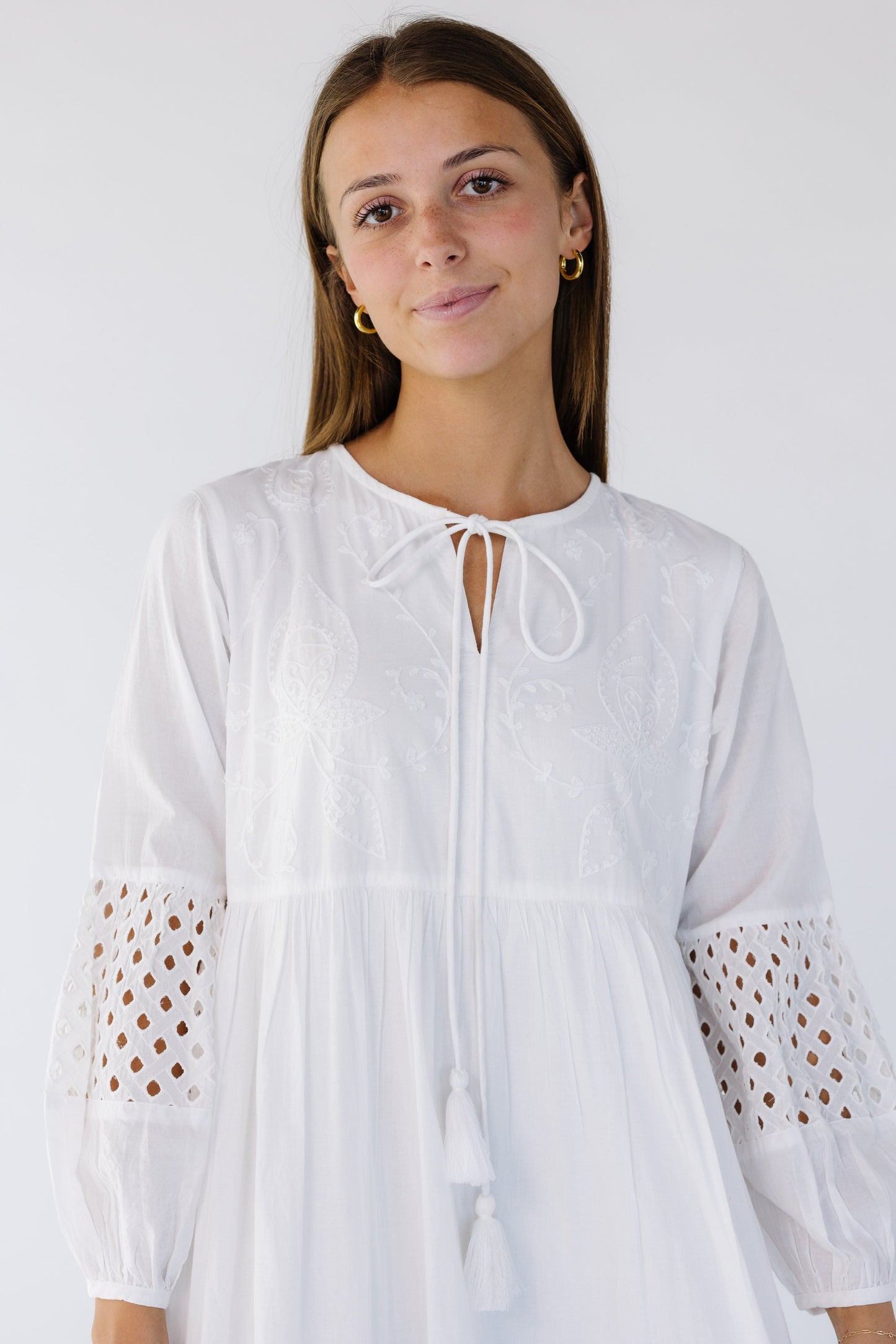 Close view of a woman wearing a white maxi dress with and embroidered bodice, round neckline with a tie closure and eyelet patterned sleeve sections.