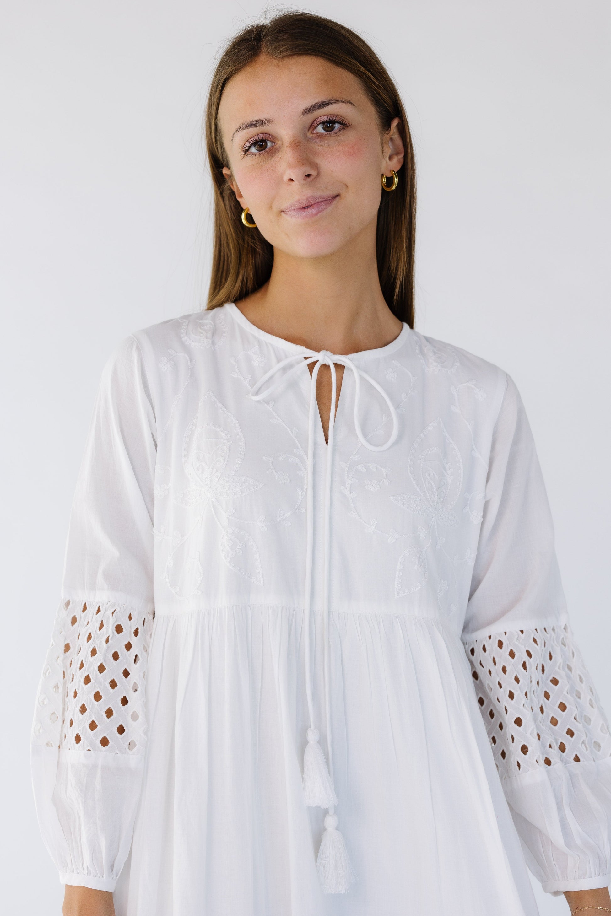 Close view of a woman wearing a white maxi dress with and embroidered bodice, round neckline with a tie closure and eyelet patterned sleeve sections.