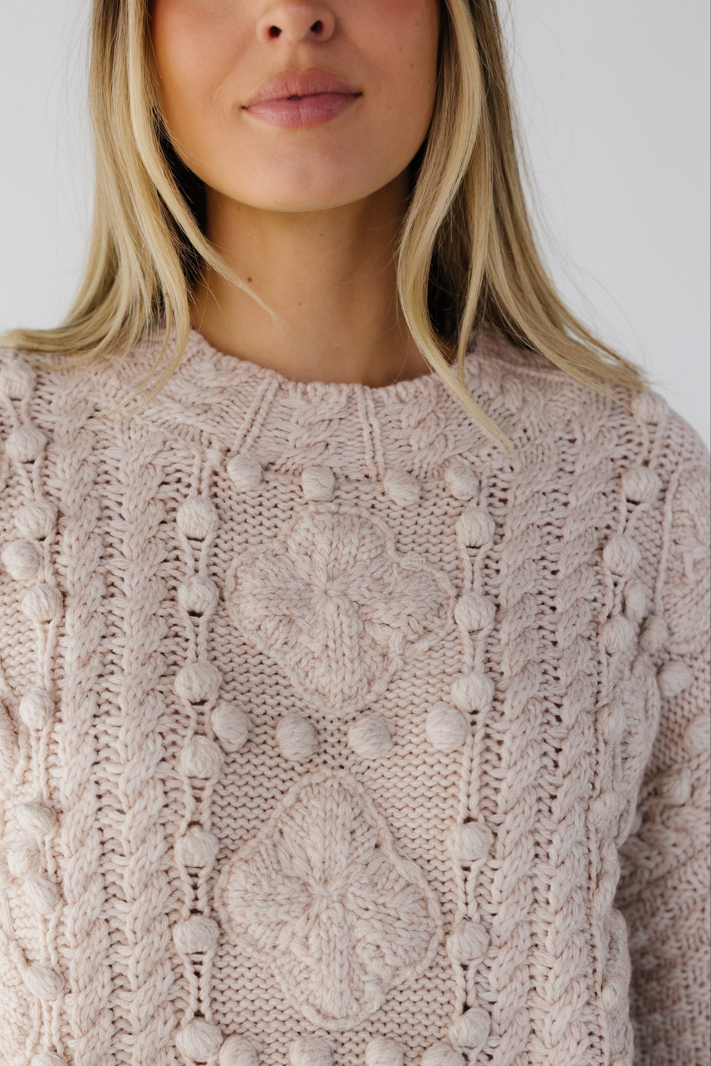 Close view of woman wearing & pom pom, cable knit & floral knit pullover sweater with a cable knit crew neckine and long sleeves.