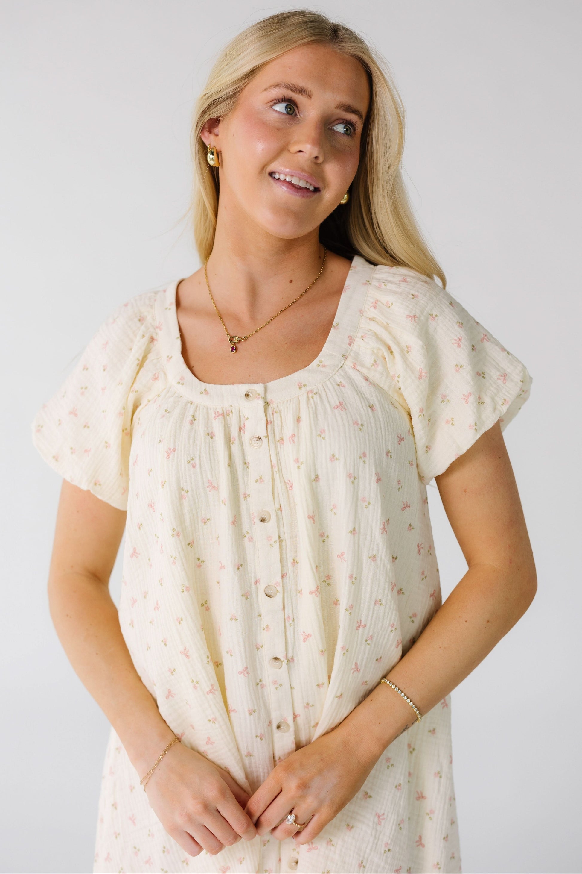 Close view of a woman wearing a mini button down dress with a scoop neckline and balloon sleeve in a cream and pink bow print.