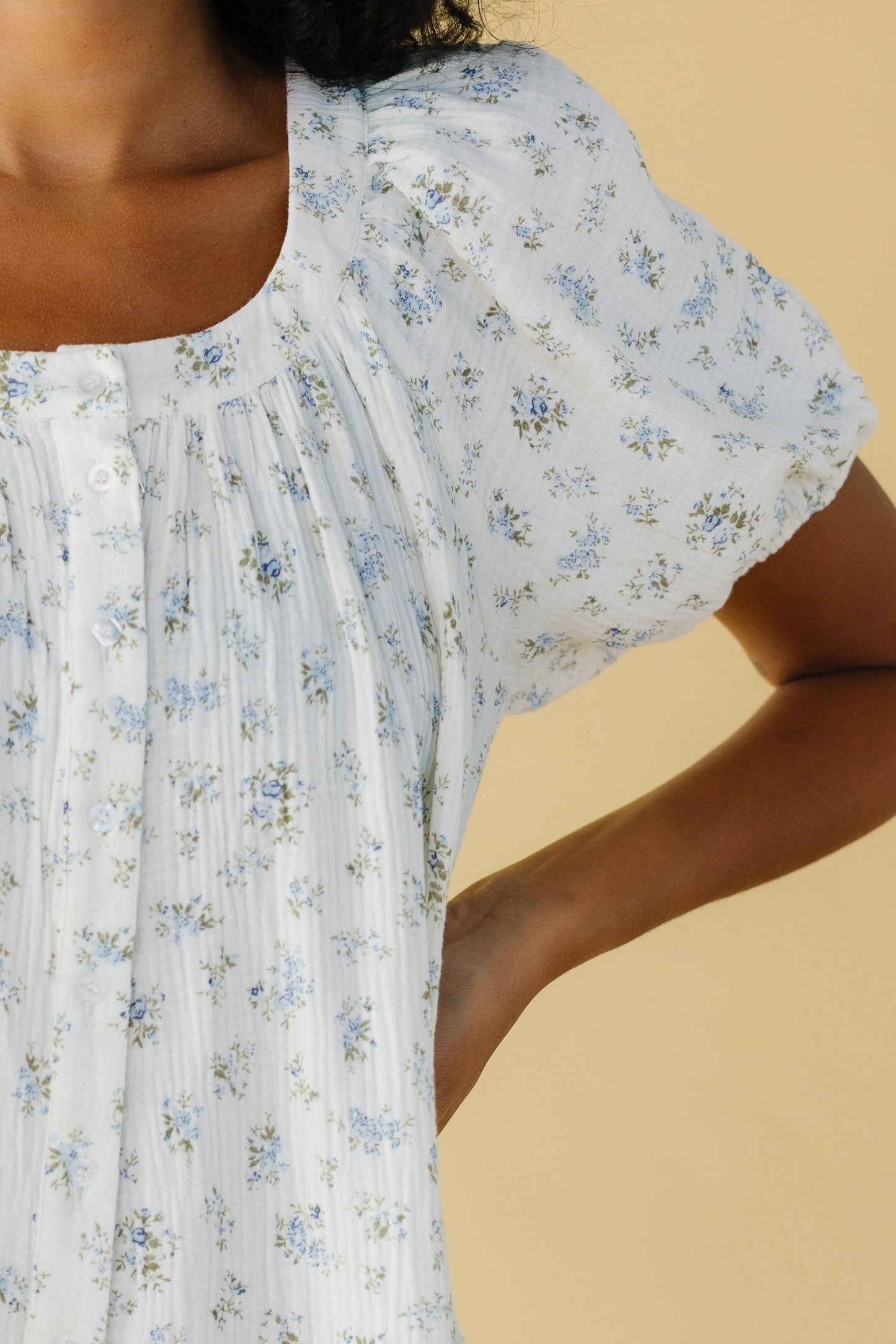 Close view of a woman wearing a white and blue floral print button down dress with a scoop neckline and large puffy sleeves.
