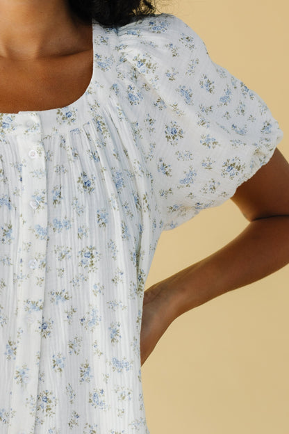 Close view of a woman wearing a white and blue floral print button down dress with a scoop neckline and large puffy sleeves.