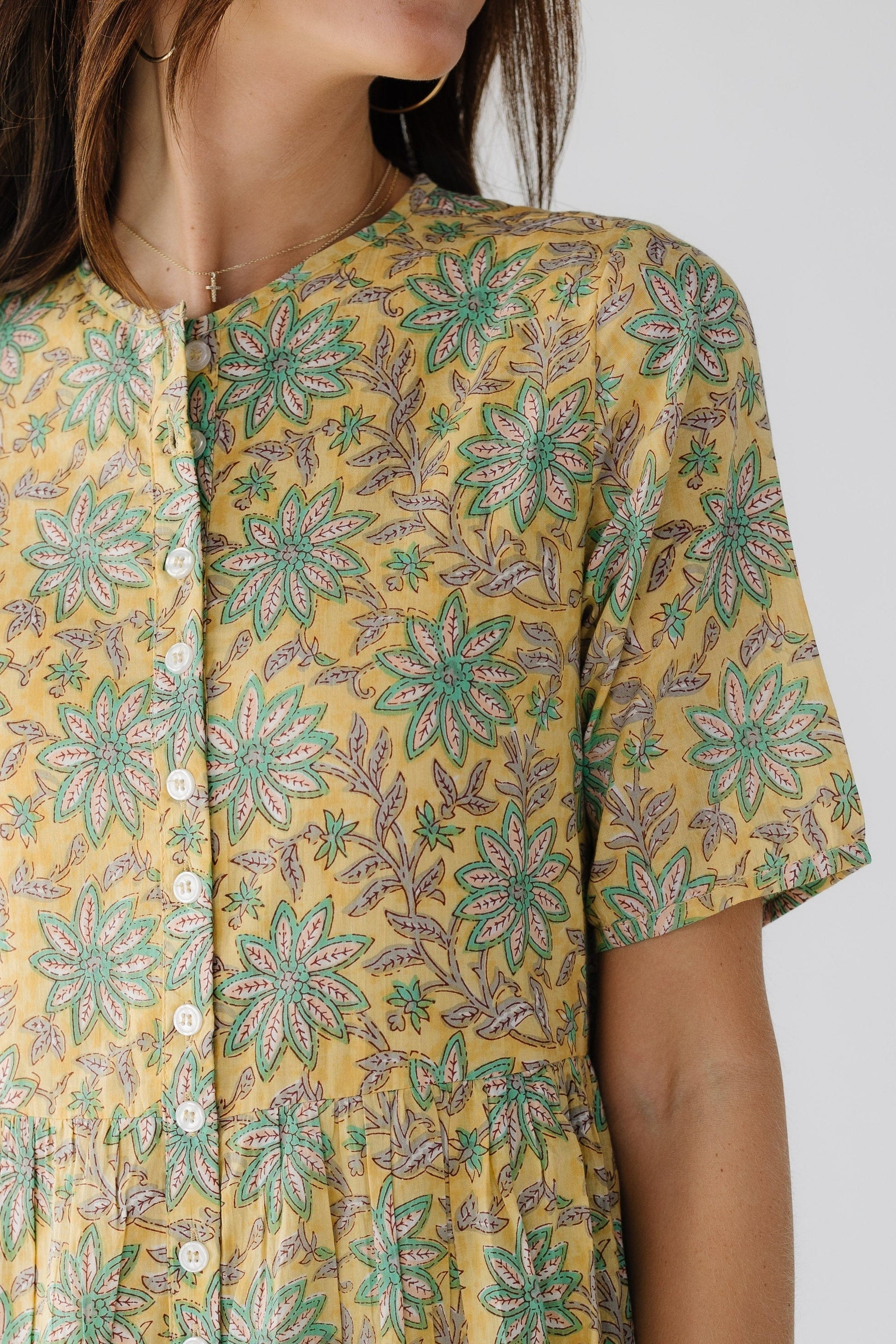 Close view of a woman wearing a yellow floral print, button down dress with short sleeves and a round neckline.