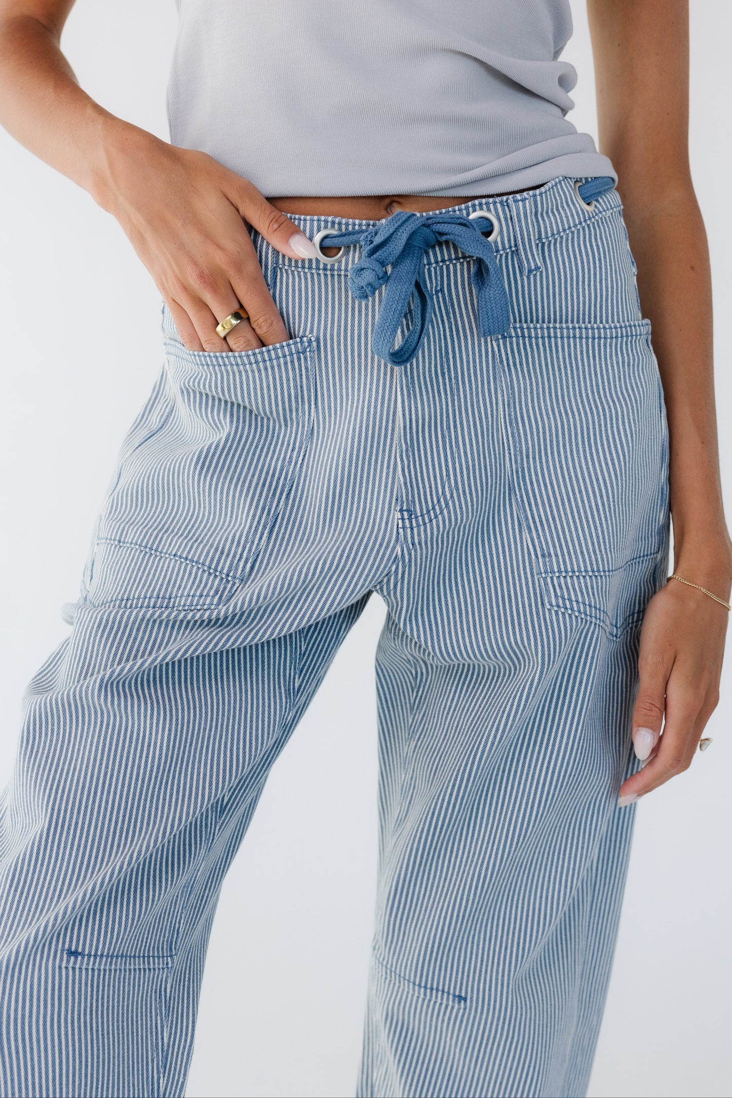 Close view of a woman wearing blue stripe barrel jeans with large front patch pockets, and large grommets in the waistband with a tie belt.
