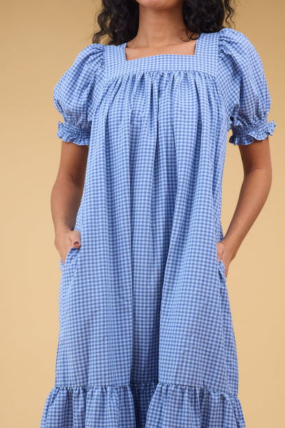 Close view of a woman wearing a blue gigngham midi dress with side pockets, sqaure neckline and short puff sleeves.