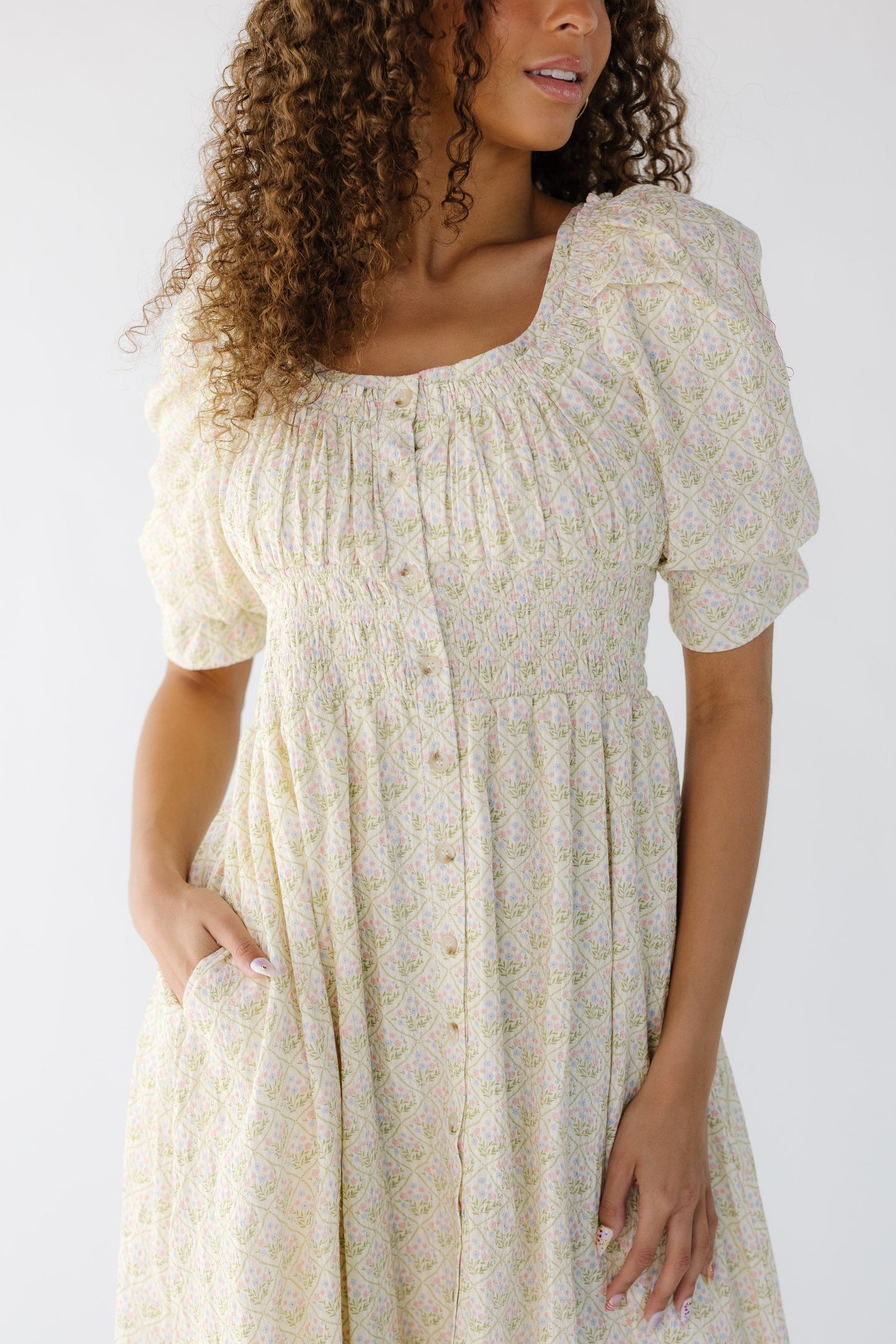 Closer view of a teenage girl wearing a  cream floral midi that buttons down the front with a wide scoop neckline, a wide smocked waist and short puff sleeves.