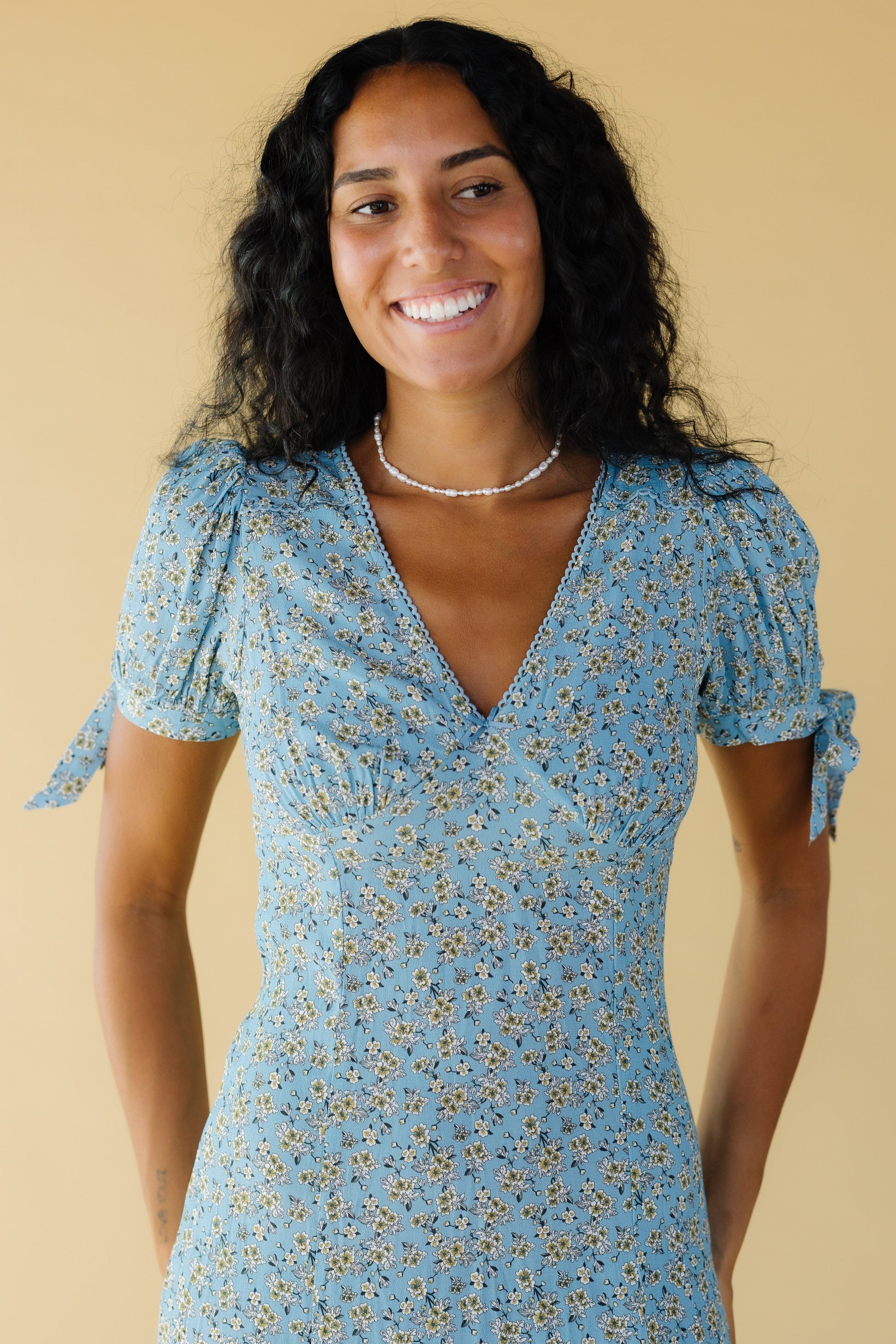 Close up view of woman wearing a light blue floral dress with a pointed empire waist, V-neckline and puff sleeves with ties.