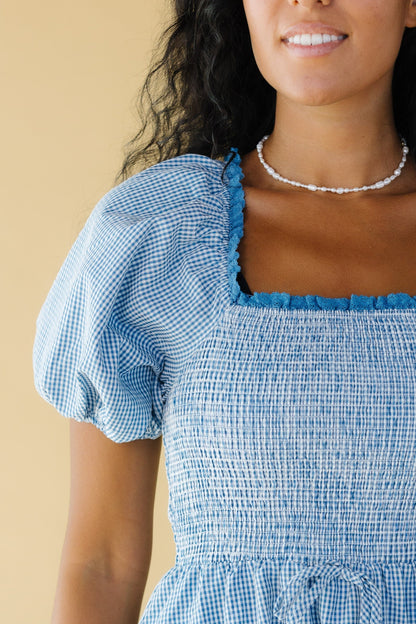 Close view of a woman wearing a blue & white gingham dress with balloon sleeves, a lace trimmed square necklace and a smocked bodice.