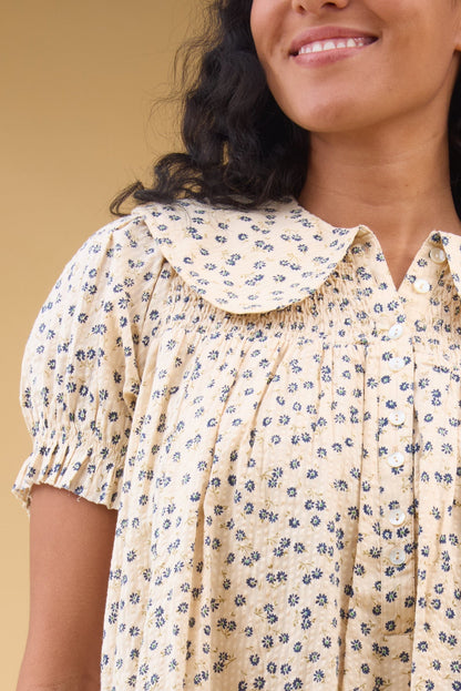 Close view of a woman wearing a cream with blue floral print dress with puff sleeves, smocked yoke and a rounded collar.