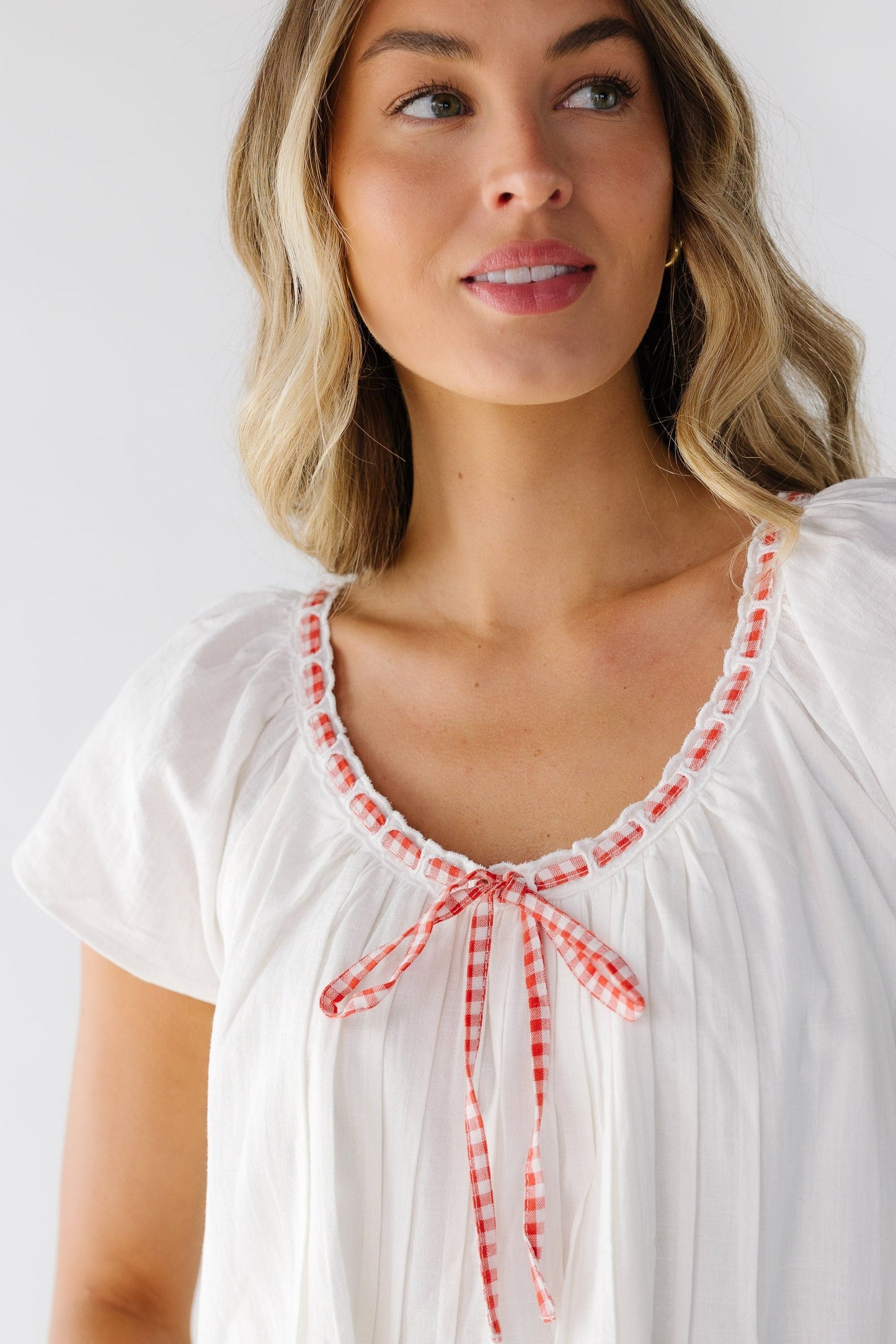 Close up view of a young woman wearing a white loose fitting dress with a scooped, eyelet lace trimmed neckline with a red & white gingham tie woven through the lace and with a tie bow in the front.