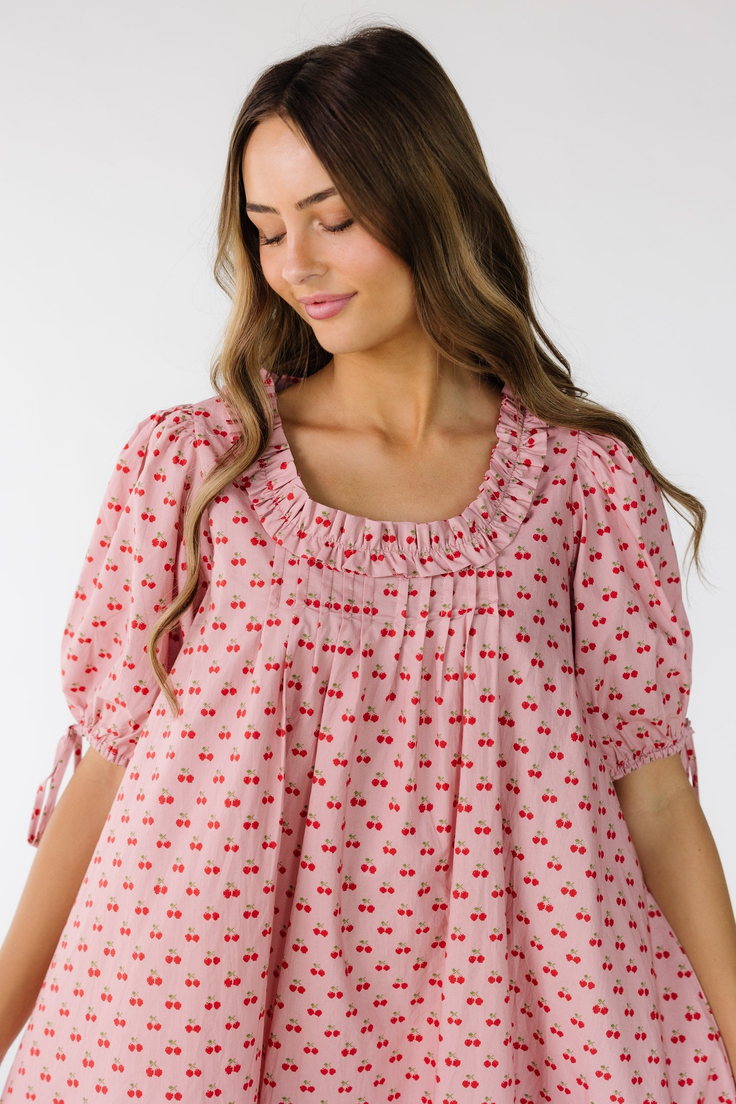 Close view of a young woman wearing a pink dress with red cherry print with a squarish neckline with pleats and ruffle details.