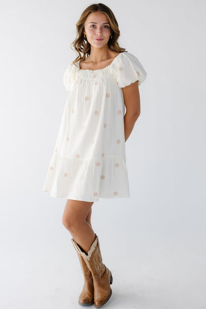 Young woman wearing an ivory short dress with pink floral embroidery, a square neckline and large puff sleeves.