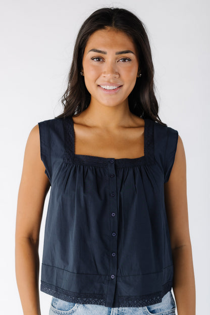 Young woman wearing a navy sleeveless, button down top with a square neckline and lace trim at the neckline and hem.