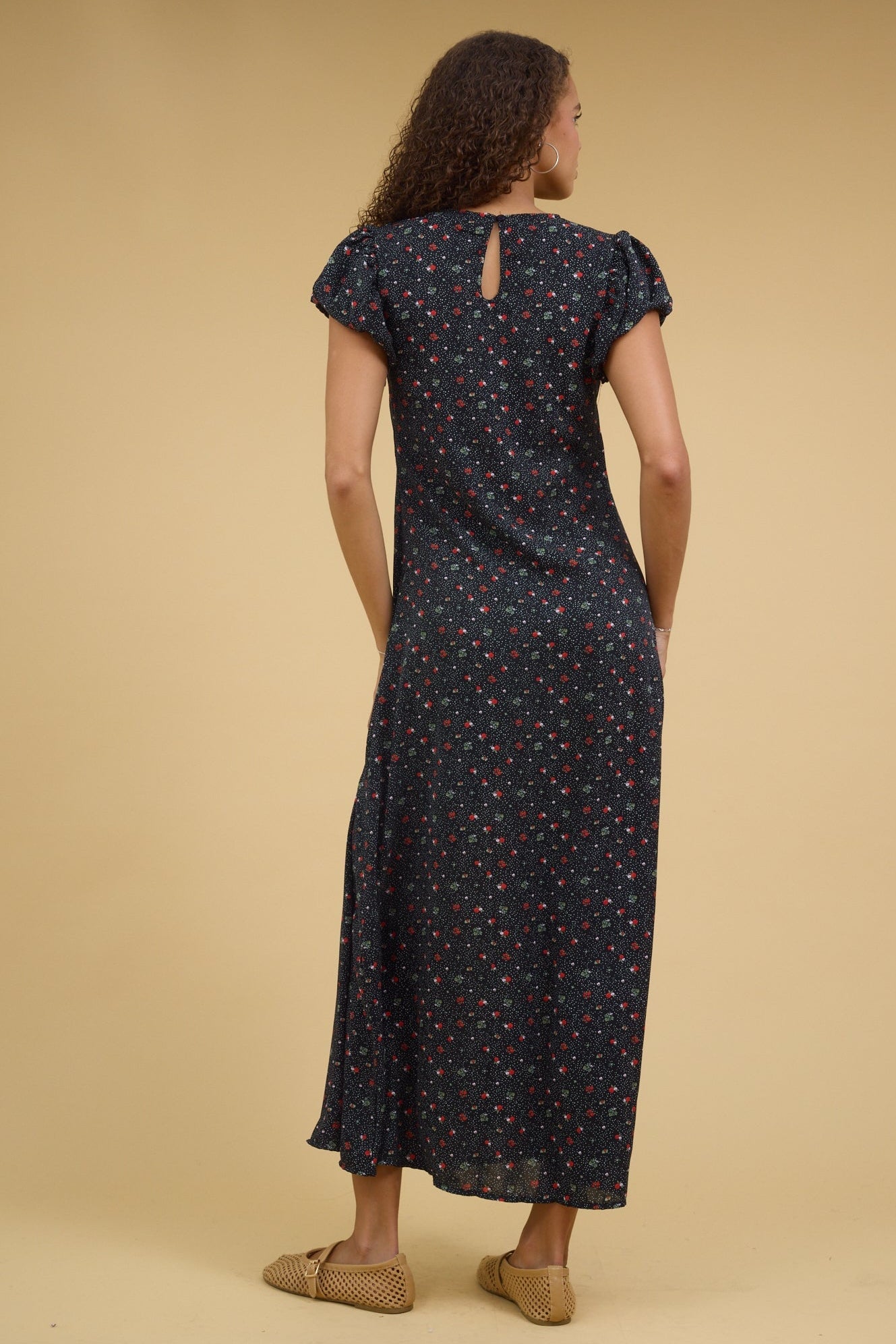 Back view of a teenage girl wearing a close fitting black floral maxi dress with gathered cap sleeves.