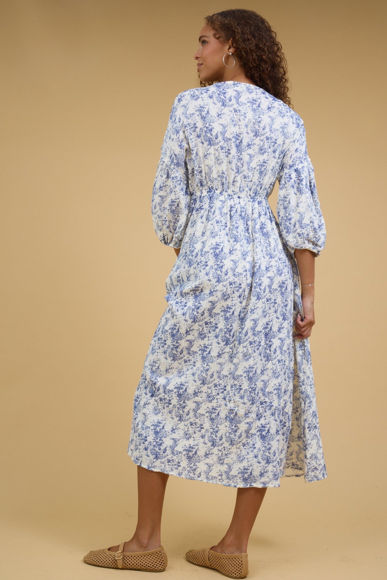 Back view of a woman wearing a white & blue print midi dress with an elastic waist and below the elbow length sleeves.