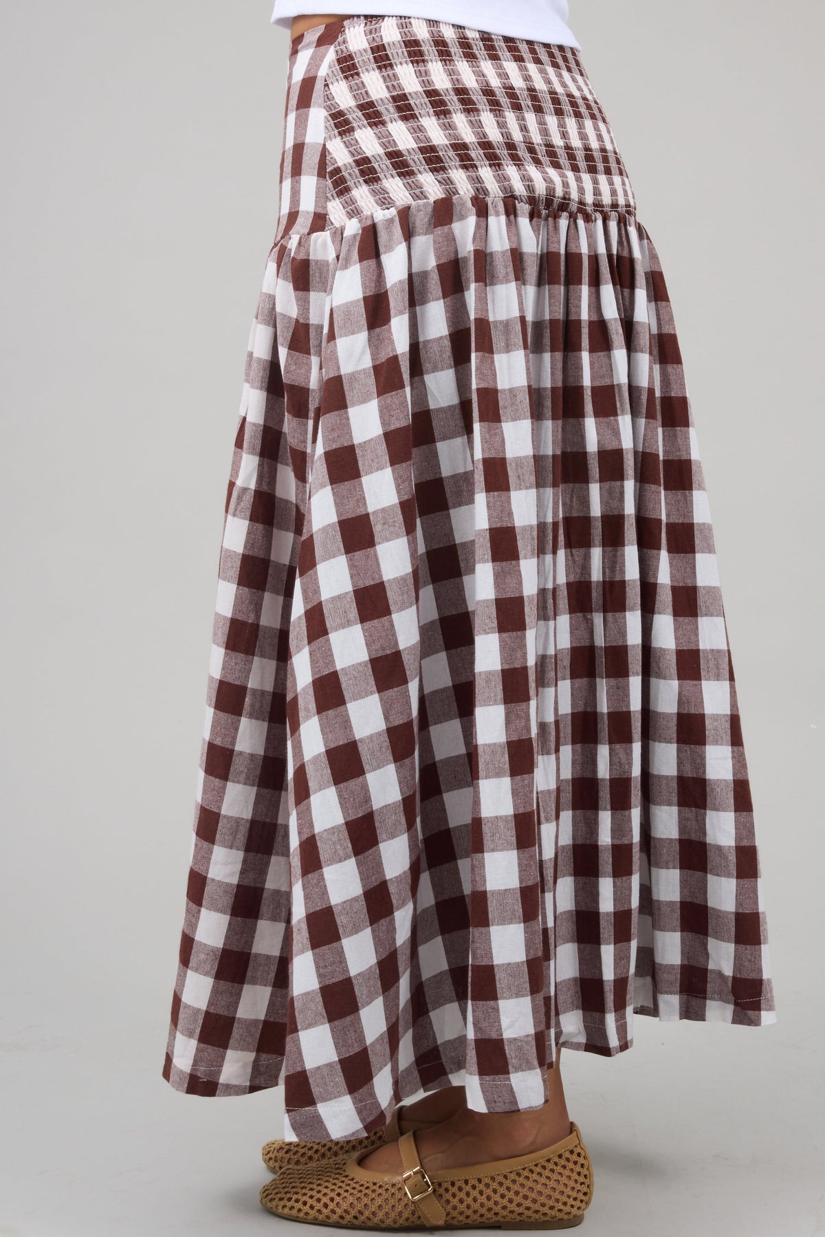 Side view of a young woman wearing a drop waist midi skirt in brown & white large gingham print with a elastic smocked back, hip panel.