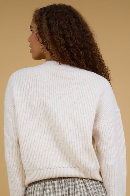Back view of a young woman wearing a cream, rib knit sweater with long sleeves.