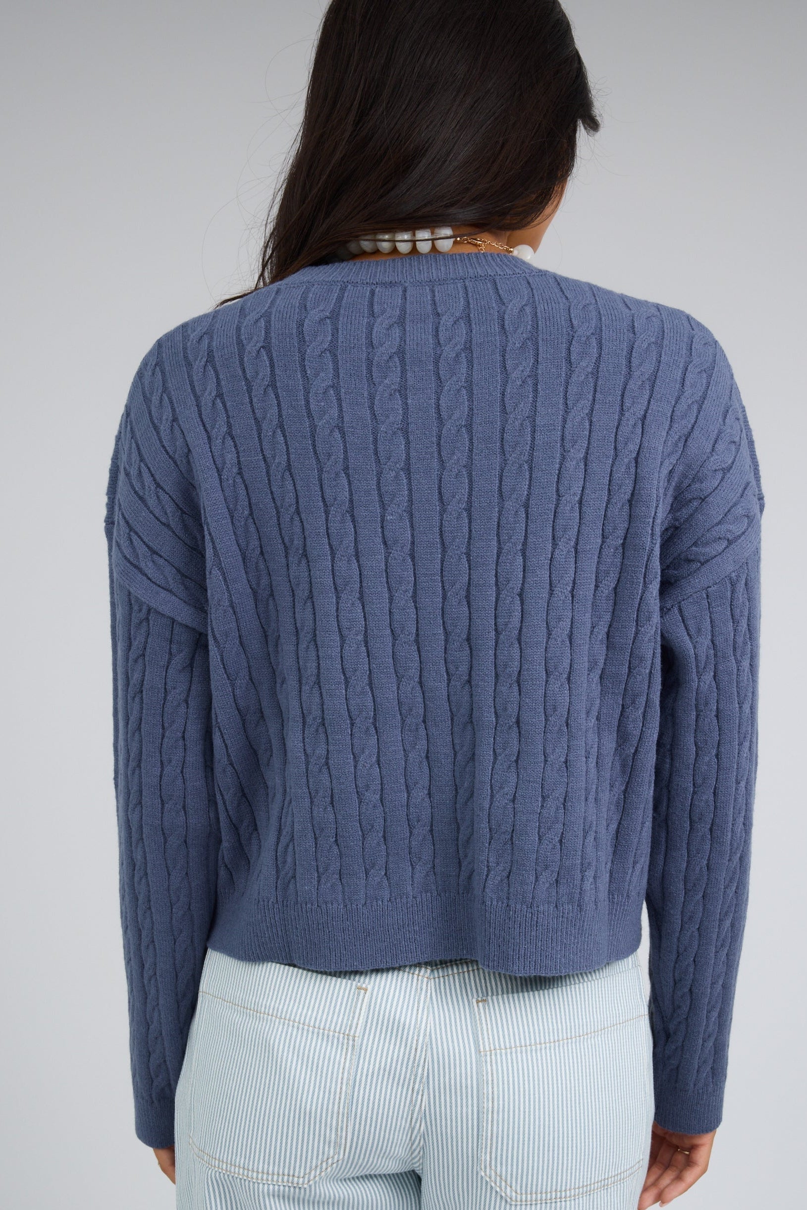 Back view of a woman wearing a blue cable knit sweater with drop shoulders and long sleeves.