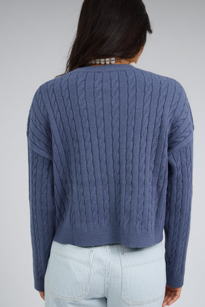 Back view of a woman wearing a blue cable knit sweater with drop shoulders and long sleeves.