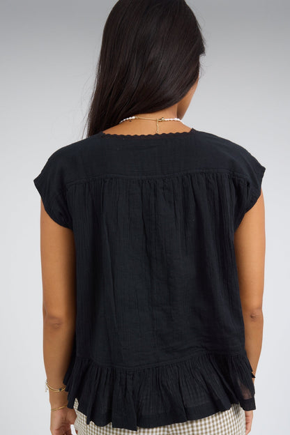 Back view of a woman wearing a loose fitting black blouse with cap sleeves and a bottom ruffle.
