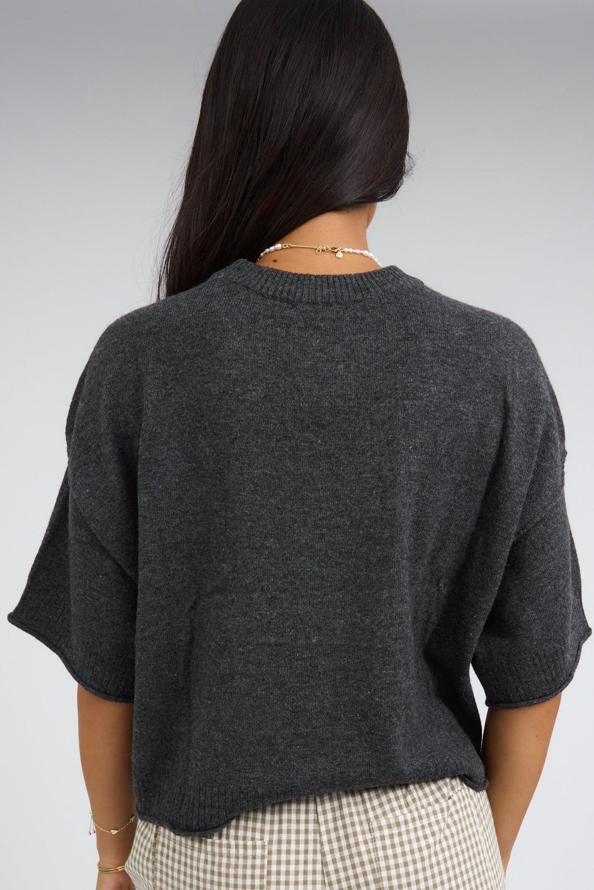 Back view of a woman wearing a charcoal loose fitting, lightweight sweater with loose elbow length sleeves and a round neckline.