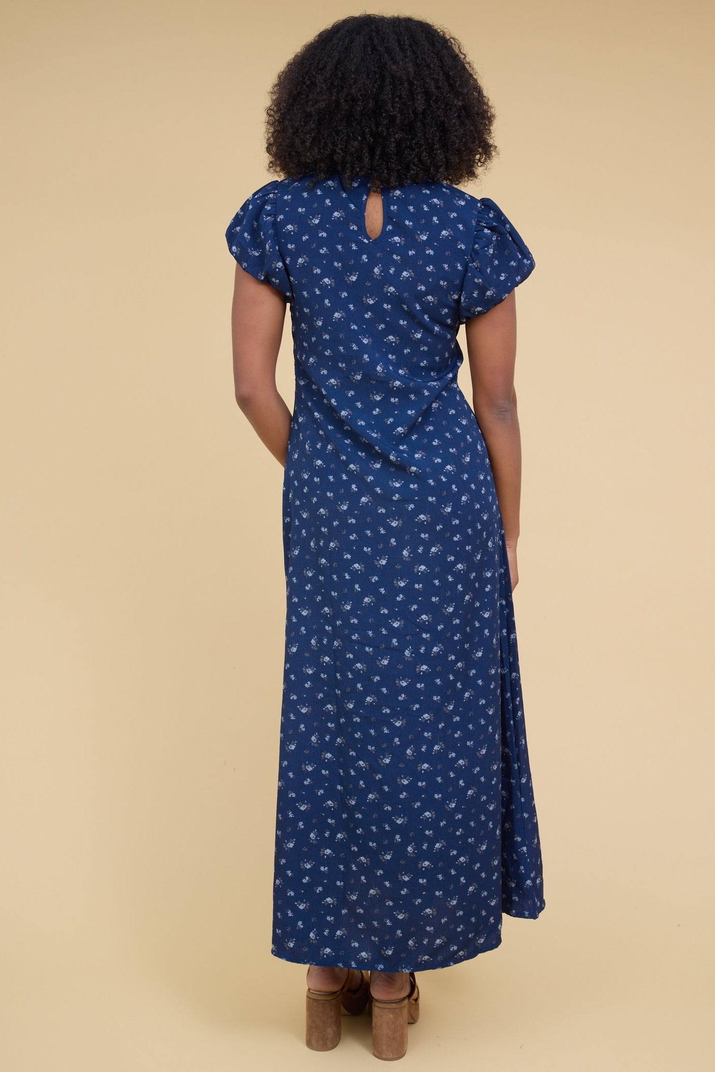 Back view of a woman wearing a close fitting blue floral maxi dress with a gathered cap sleeve.