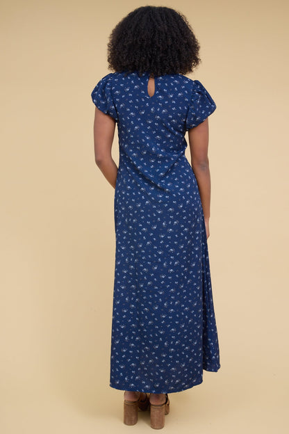 Back view of a woman wearing a close fitting blue floral maxi dress with a gathered cap sleeve.