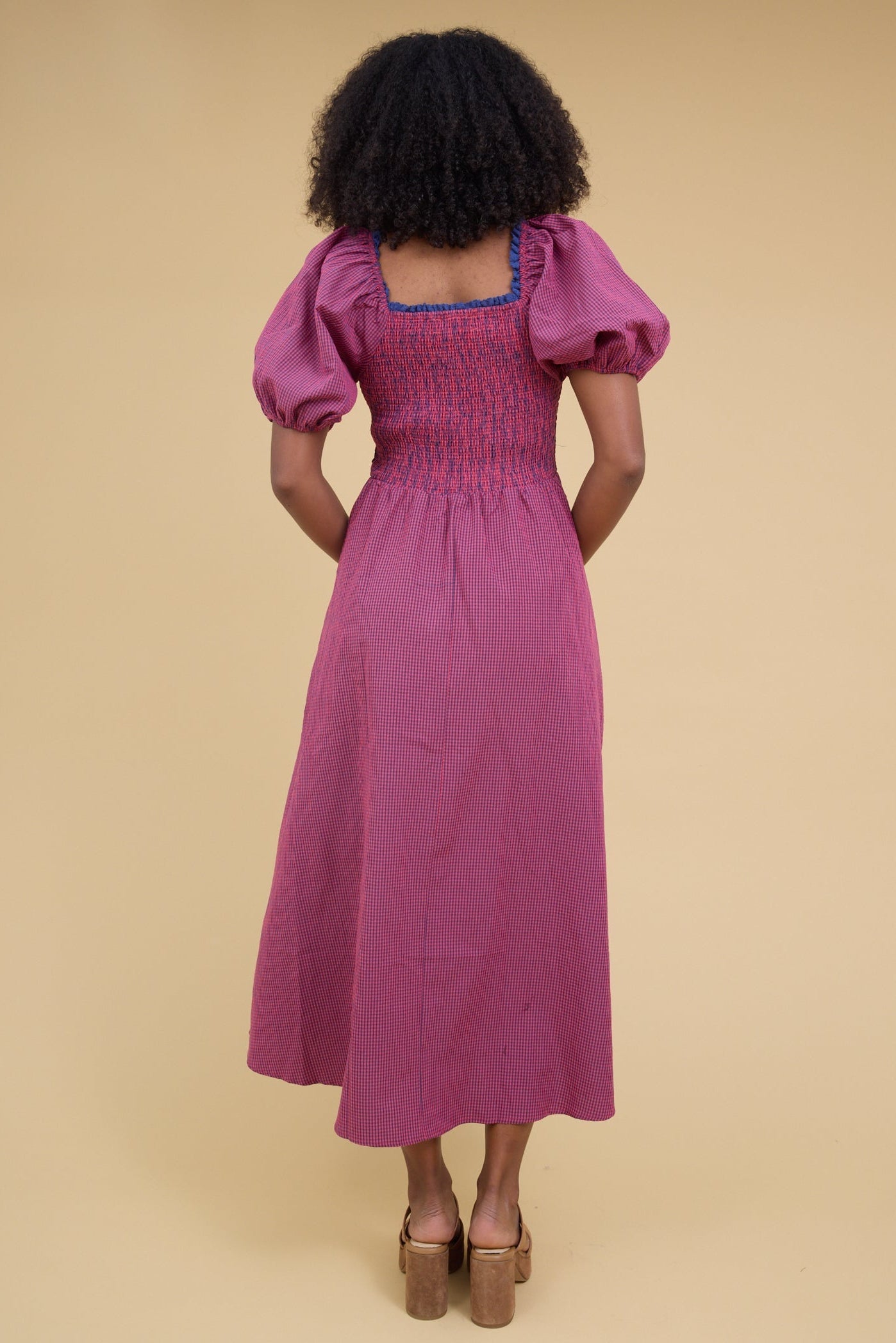 Back view of a woman wearing a red & blue gingham midi dress with a square neckline, balloon sleeves and a smocked bodice.