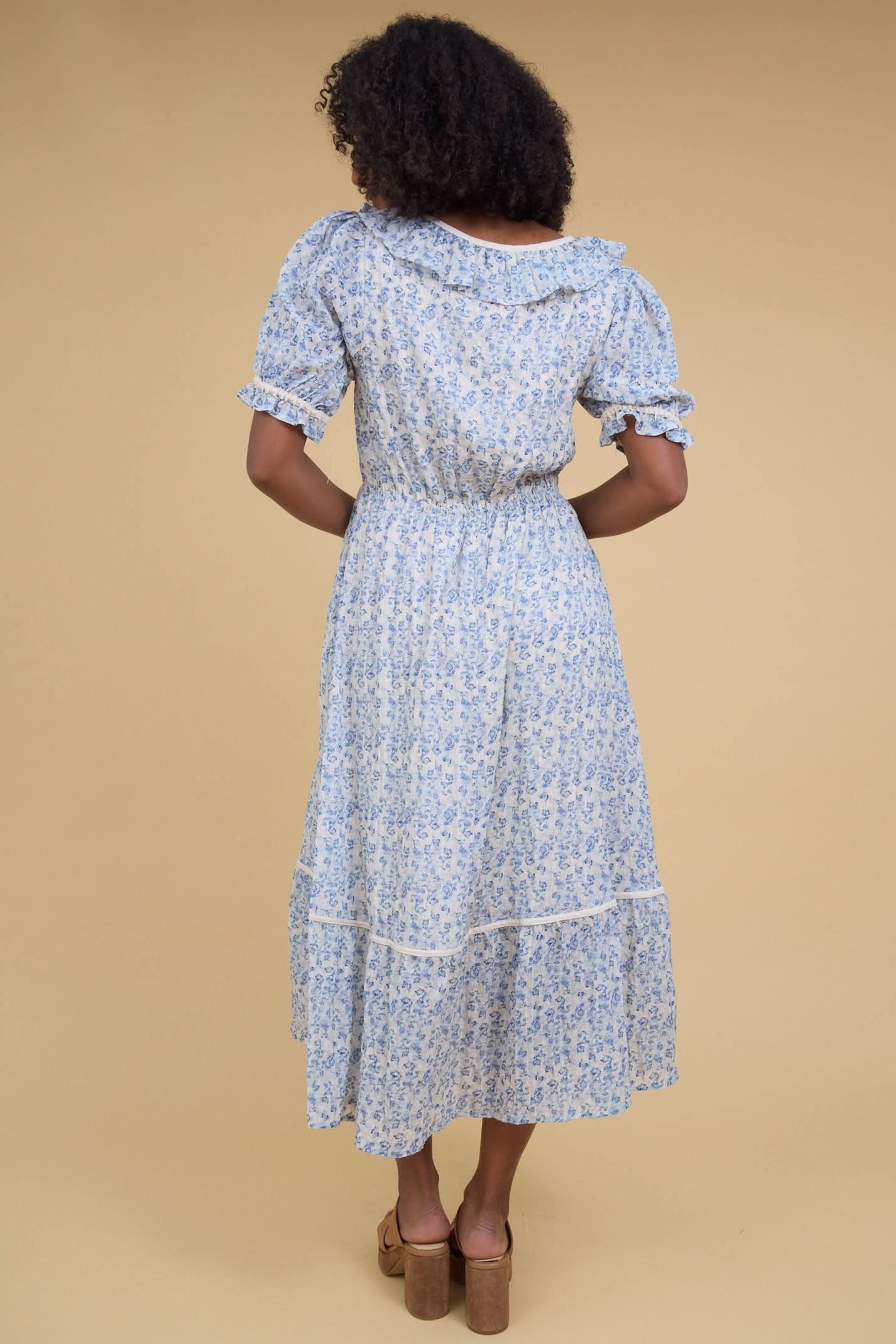 Back view of a woman wearing a white and blue floral print with a ruffled neckline, puff sleeves and a full midi skirt.