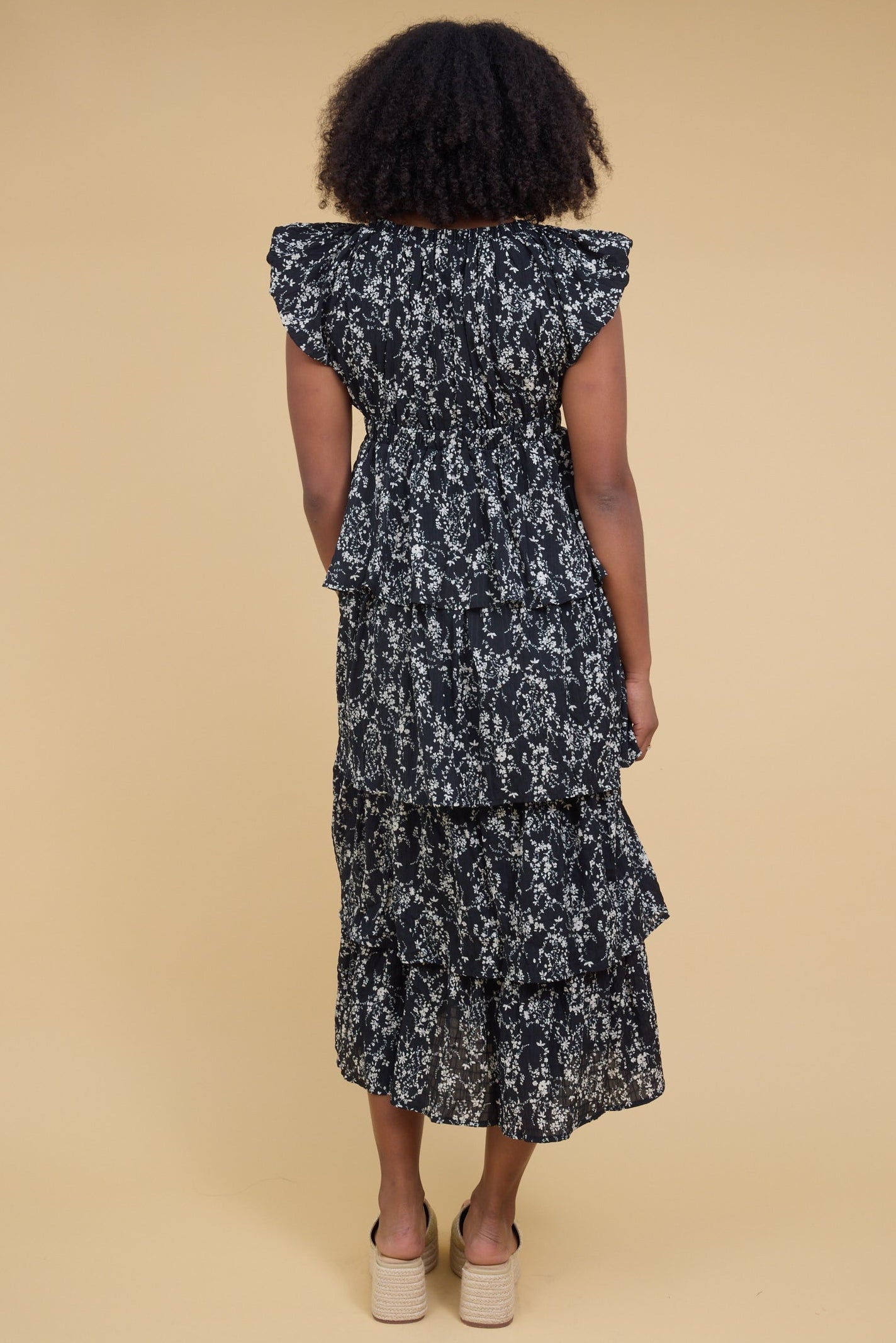Back view of a woman wearing a black & white floral print midi dress with loose layered tier skirt with an empire waist and short gathered cap sleeves.