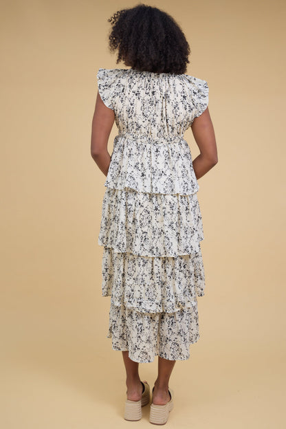 Back view of a woman wearing an off white & black floral print midi dress with loose layered tiers and short sleeves.