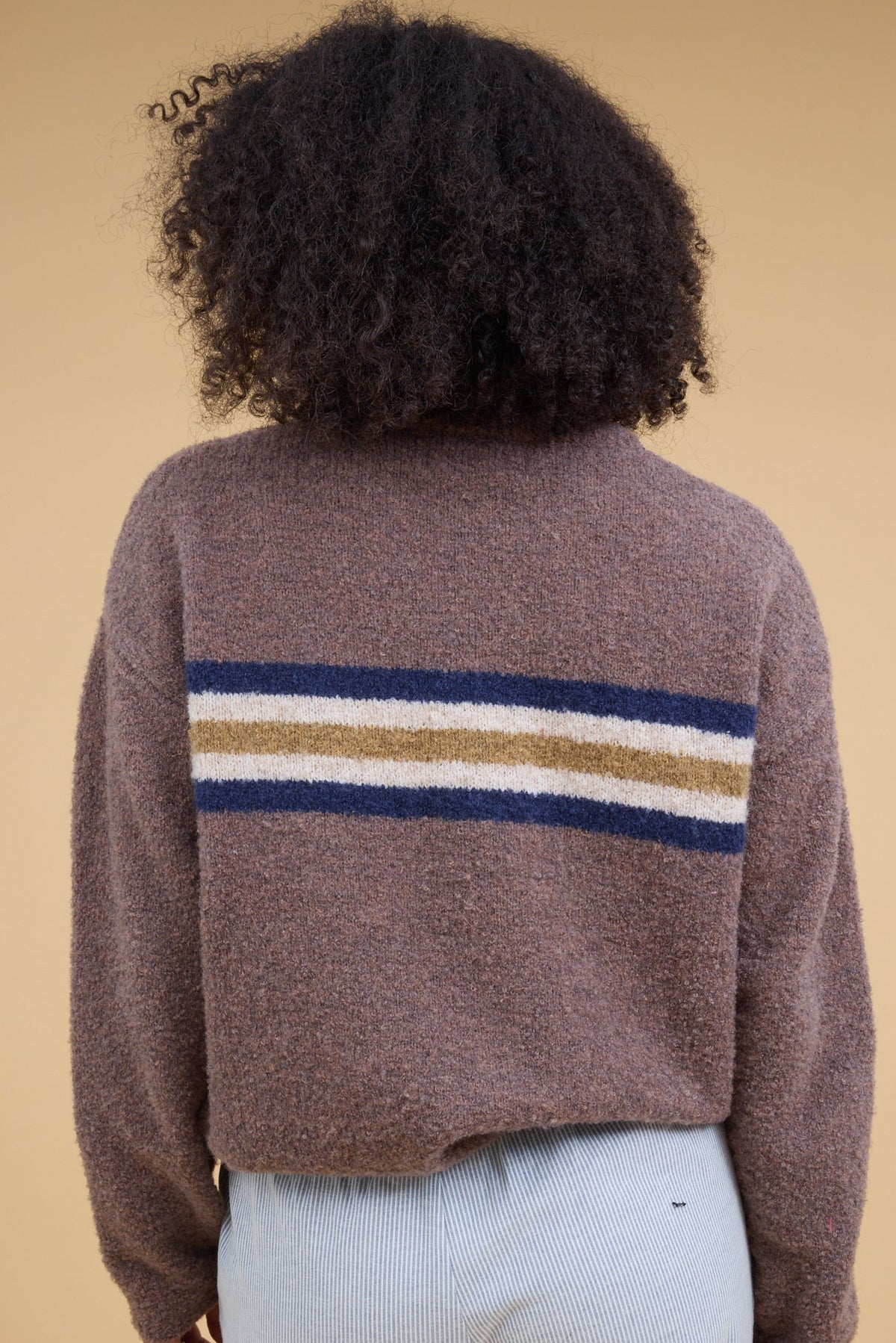 A back view of a young woman wearing a light brown sweater with long sleeves and a singe wide chest stripe in blue, white  gold.