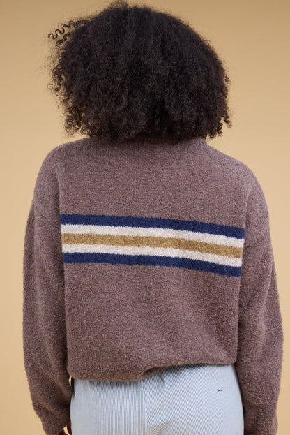 A back view of a young woman wearing a light brown sweater with long sleeves and a singe wide chest stripe in blue, white  gold.