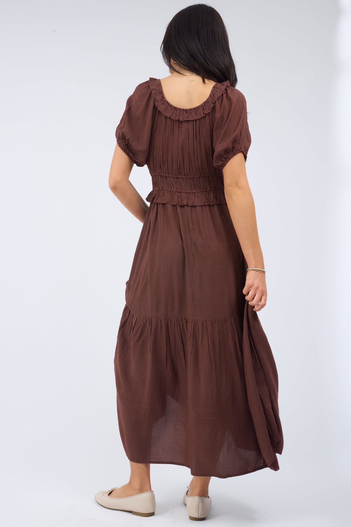 Back view of a woman wearing a loose fitting midi dress with a back ruffled scoop neckline with large puff sleeves and a wide waistband.