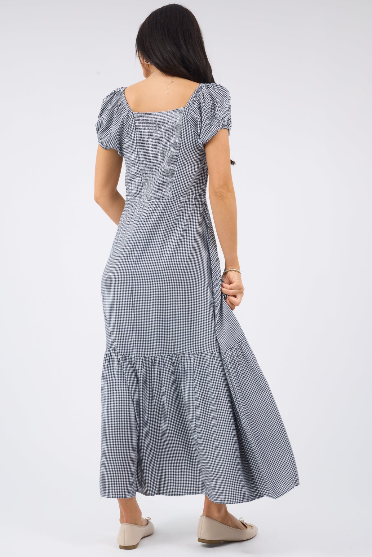 Back view of a woman wearing a navy gingham midi-maxi dress with a wide bottom ruffle, short puff sleeves and an elastic smocked back bodice panel.