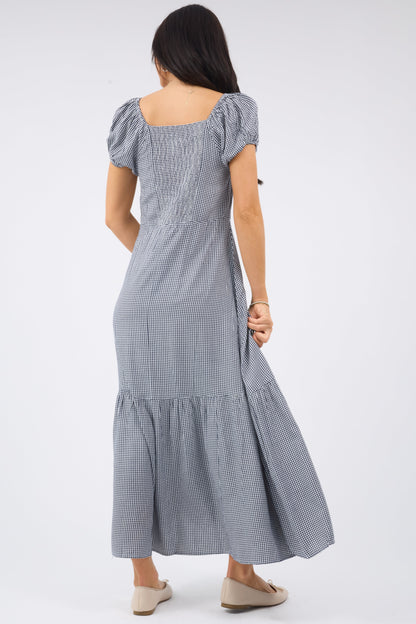 Back view of a woman wearing a navy gingham midi-maxi dress with a wide bottom ruffle, short puff sleeves and an elastic smocked back bodice panel.