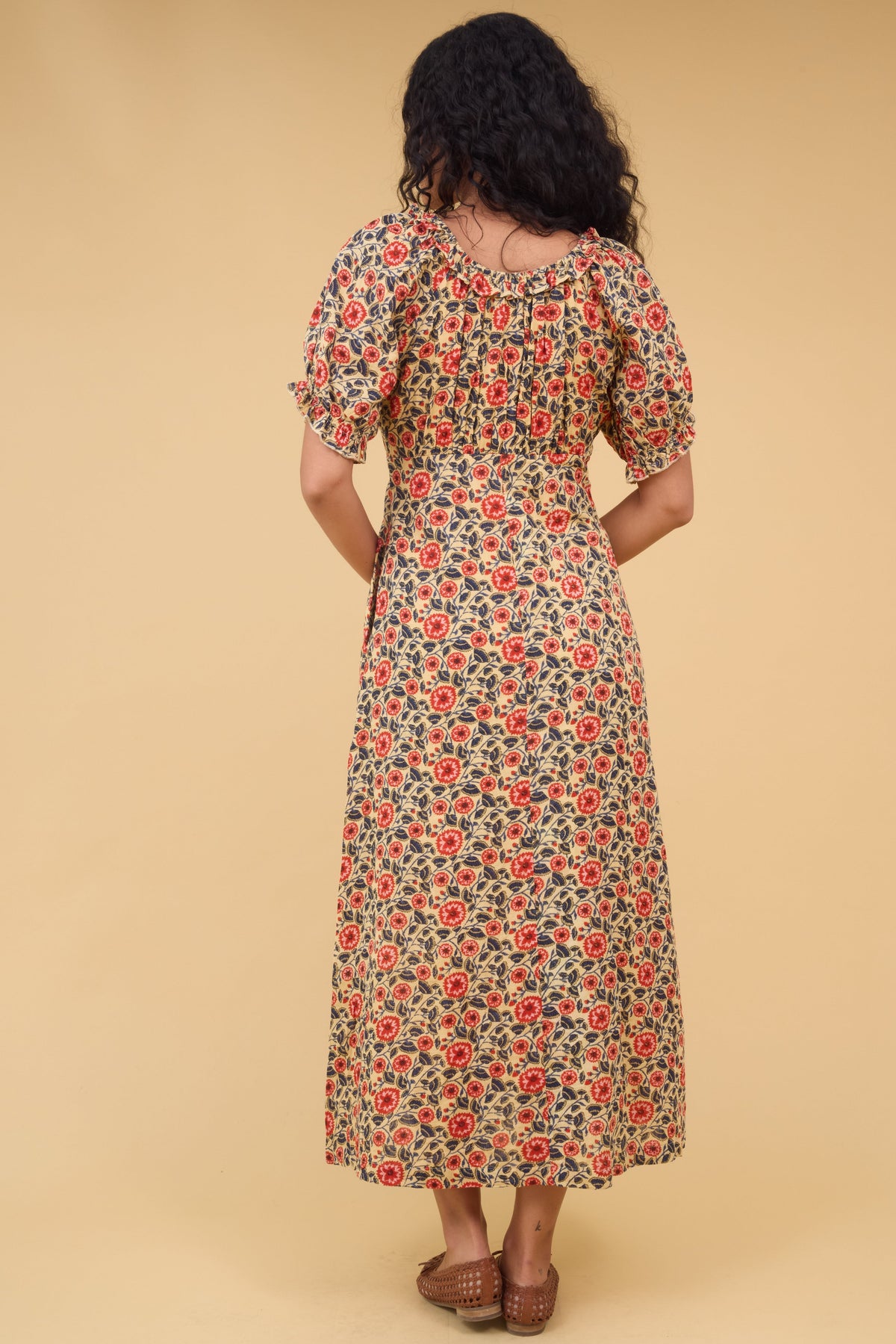Back view of a woman wearing a floral print midi dress with a gathered bodice, short sleeves and a ruffled neckline.