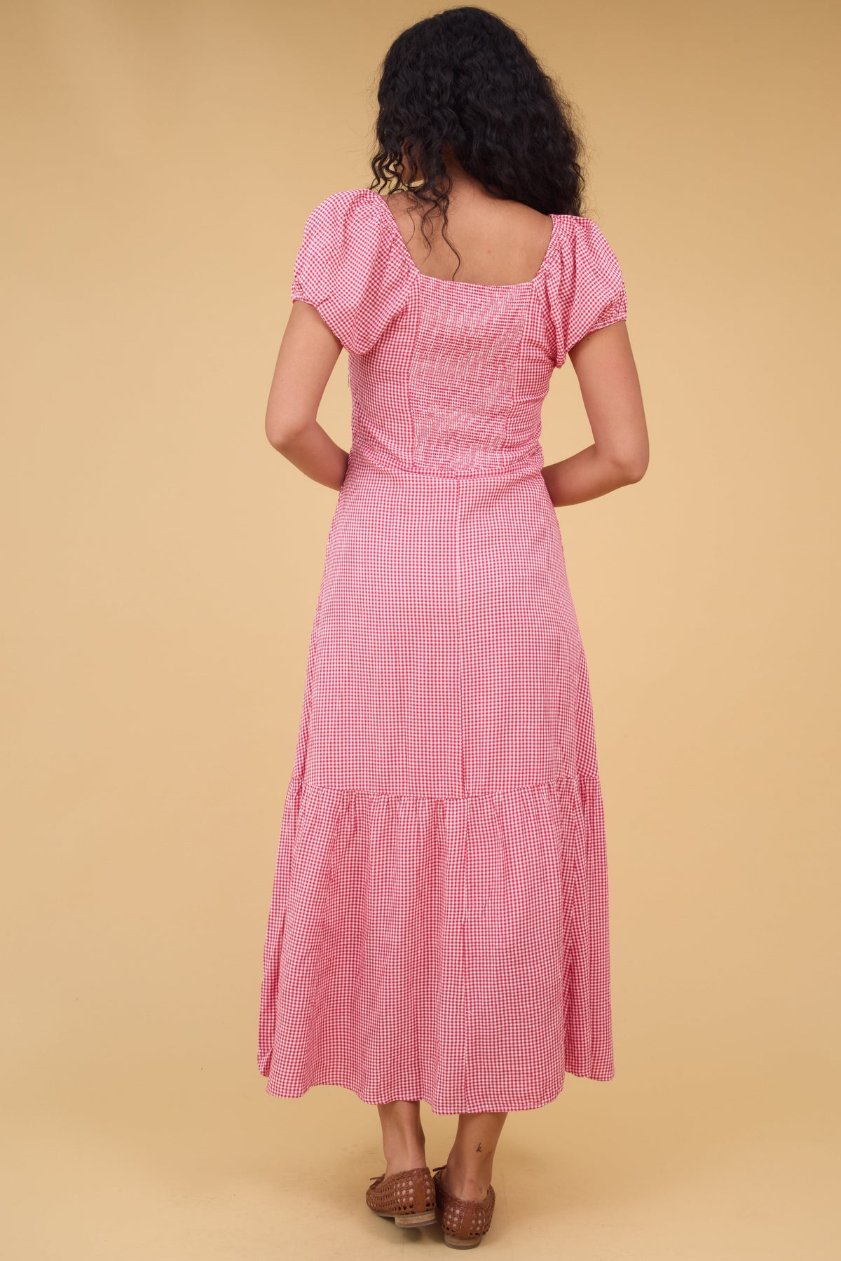 Back view of a woman wearing a pink gingham dress wtih a square neckline short puff sleeves and a elastic smocked back bodice panel.