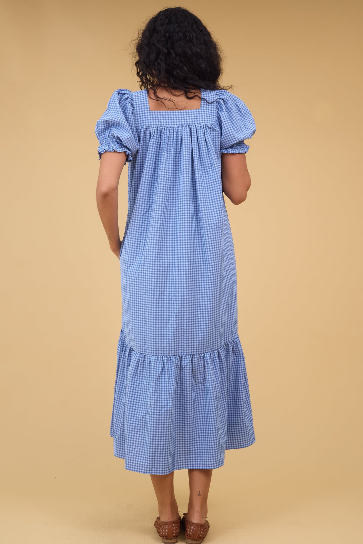 Back view of a woman wearing a loose fitting blue gingham midi dress with a square neckline.