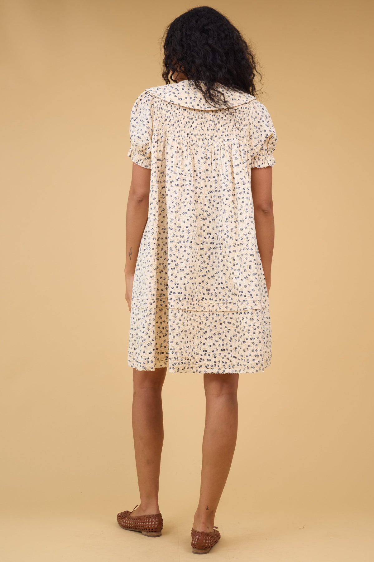 Back view of a woman wearing a short cream dress with a smocked yoke, short sleeves and a wide collar.