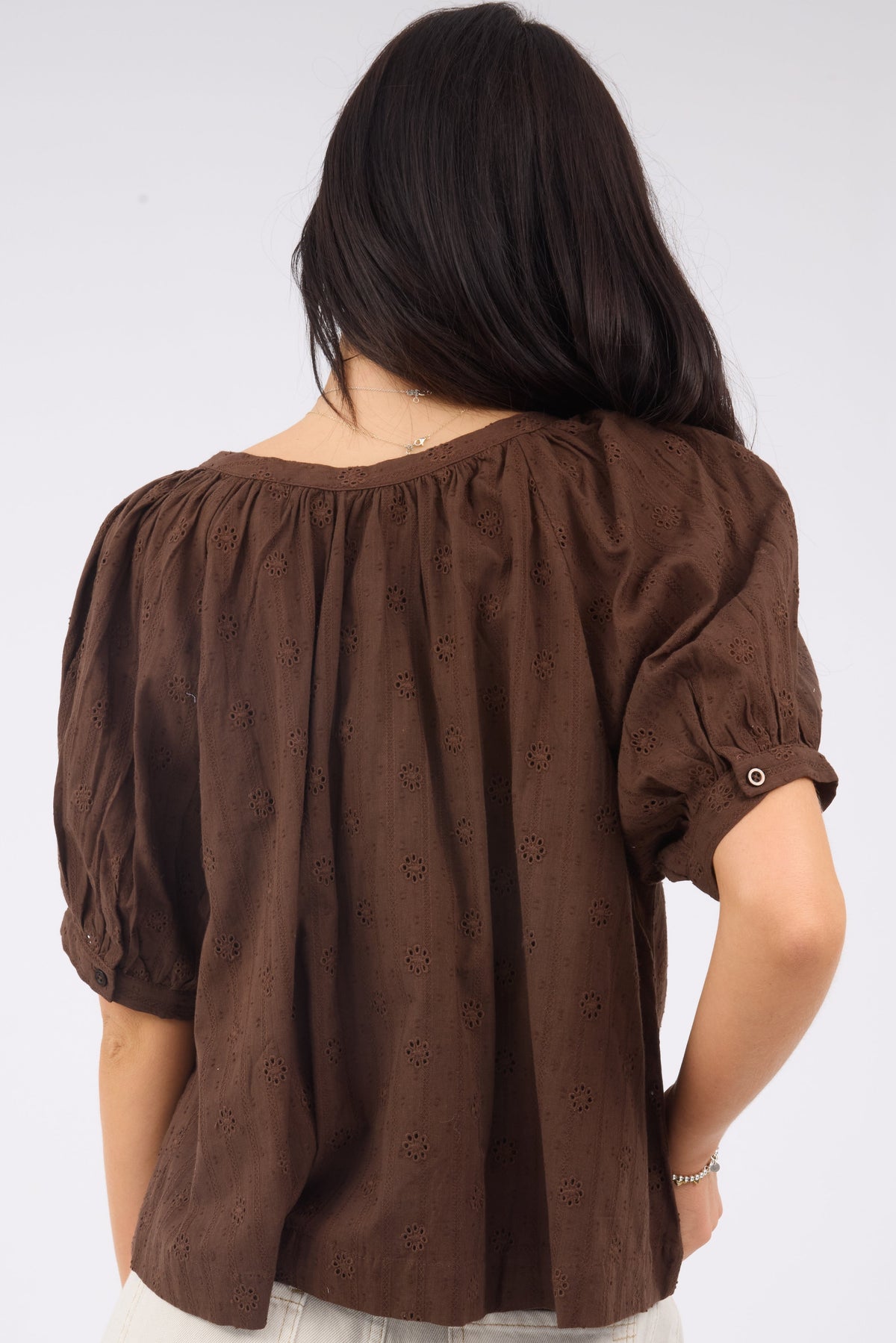 A back view of a woman wearing a loose fitting brown eyelet blouse with shrot puff sleeves.