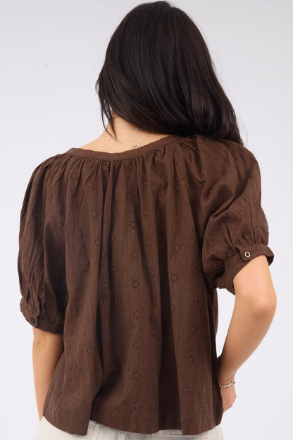 A back view of a woman wearing a loose fitting brown eyelet blouse with shrot puff sleeves.