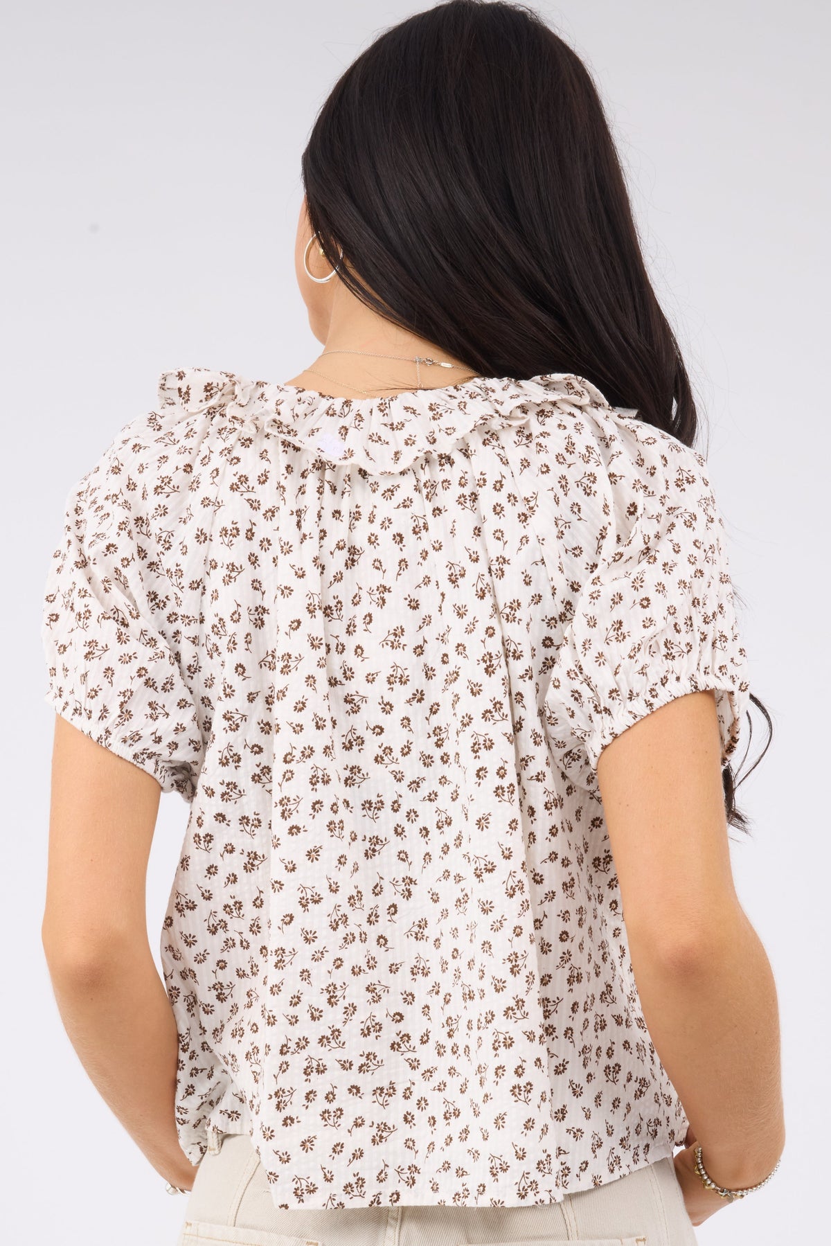 A back view of a woman wearing a loose fitting white & brown floral print blouse with a ruffle collar and short puff sleeves.