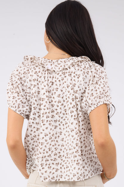 A back view of a woman wearing a loose fitting white & brown floral print blouse with a ruffle collar and short puff sleeves.