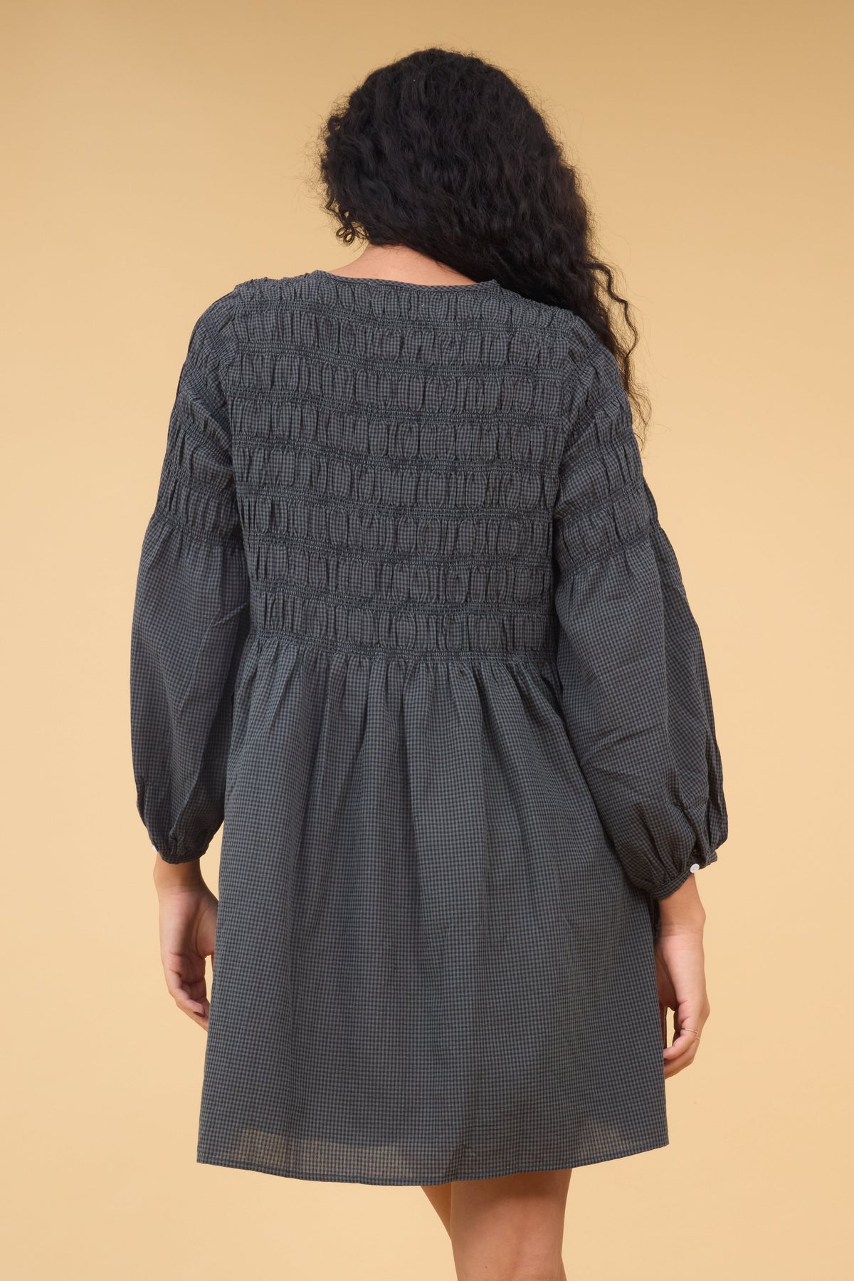 Back view of a woman wearing a loose fitting gingham dress in dark grey gingham with long sleeves and spaced out smocking stripes.