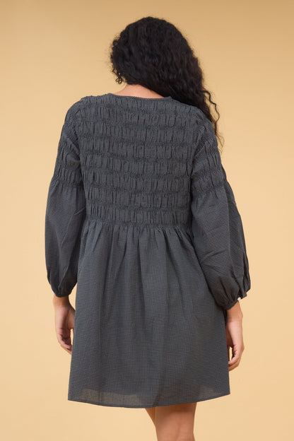 Back view of a woman wearing a loose fitting gingham dress in dark grey gingham with long sleeves and spaced out smocking stripes.