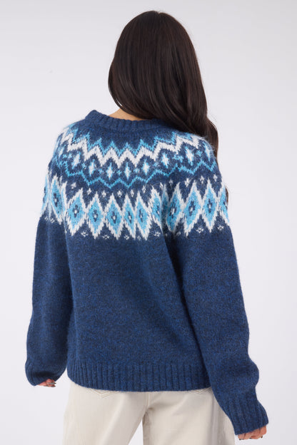A back view of a young woman wearing a navy & blue nordic sweater with a wide crew neckline and long sleeves.