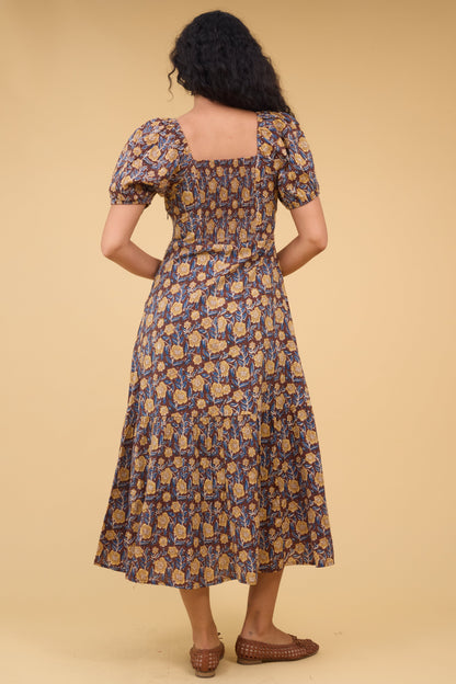 Back view of a woman wearing a floral midi dress with a wide bottom ruffle, puff sleeves and a back elastic smocked panel with a square neckline. 