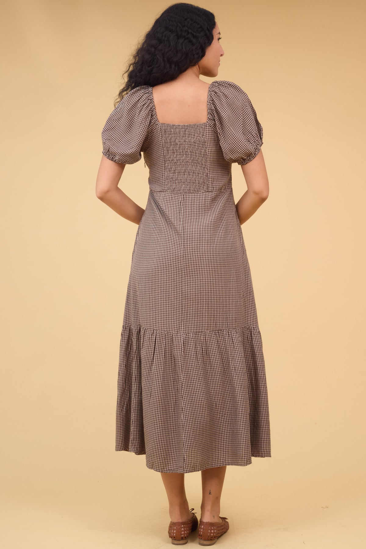 Back view of a women wearing a brown gingham midi dress with a wide bottom ruffle, elastic back bodice panel and puff sleeves.