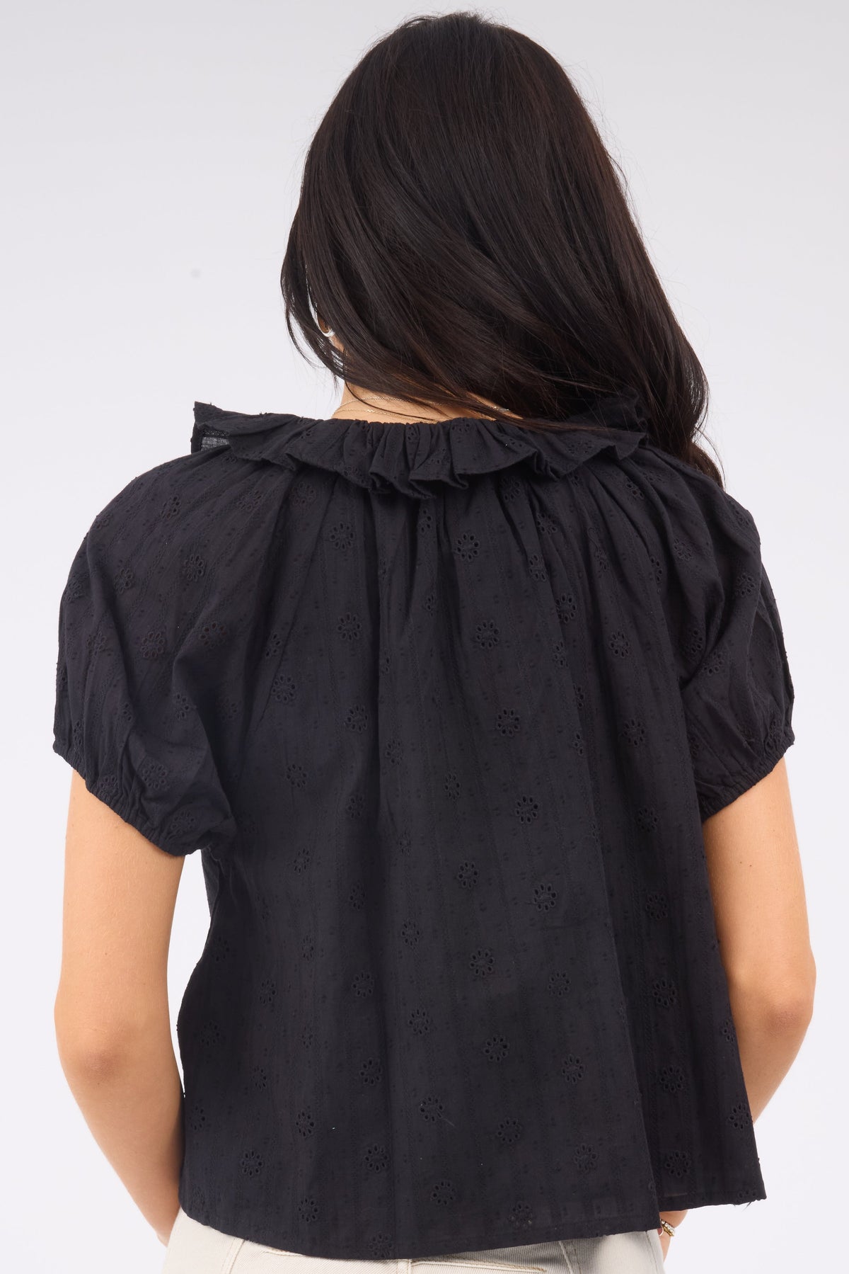 A back view of a young woman wearing a loose fitting black eyelet blouse with a ruffle neckline nad short puff sleeves.